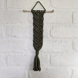 May include: Olive green macrame wall hanging. The decorative piece features a woven design with a chevron pattern, suspended from a natural wooden dowel. A tassel hangs from the bottom. The background is a white brick wall.