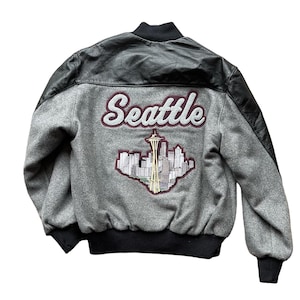 Könnte beinhalten: Eine graue und schwarze Collegejacke mit einem Lederärmel. Die Rückseite der Jacke zeigt einen gestickten Aufnäher mit dem Wort "Seattle" und einer Darstellung der Skyline der Stadt.
