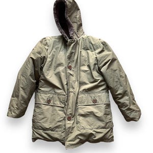 Puede incluir: Una chaqueta verde con capucha y forro de borreguillo marrón. La chaqueta tiene cierre de botones, dos bolsillos grandes y una capucha con cordón.