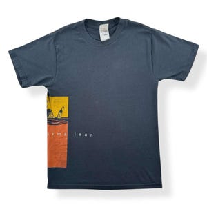 May include: Dark gray t-shirt with a rectangular graphic on the left side. The graphic features yellow and orange colors with a silhouette design. The text "orma jean" is printed in white.