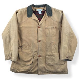 May include: Tan canvas jacket with a dark green collar and a red and black plaid lining. The jacket has four front pockets, button closures, and the L.L.Bean logo on the collar. A classic, durable outerwear piece.