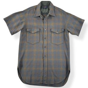 May include: A short-sleeved, button-down shirt in a gray and gold plaid pattern. The shirt has two chest pockets with button closures and a classic collar. The fabric appears to be a medium-weight material, suitable for cooler weather.