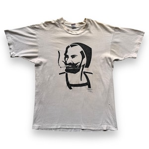 May include: A light beige t-shirt featuring a black graphic of a man smoking a cigarette. The graphic is a stylized portrait with the text "FASHION VICTIMS" printed below. The shirt has short sleeves and a crew neck.