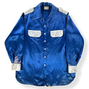 May include: A long-sleeved button-down shirt in a vibrant royal blue satin fabric. The shirt features contrasting white satin panels on the shoulder epaulets, two chest pockets with flaps, and wide cuffs. It has a pointed collar and white buttons.