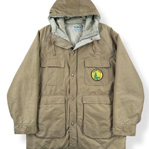 Może przedstawiać: Beżowa kurtka z kapturem z wieloma kieszeniami i naszywką z napisem "Hickory Grove Touring Co.". Kurtka ma zamek błyskawiczny i zapięcie na guziki oraz jasną podszewkę. Kurtka jest wykonana z wytrzymałego materiału.