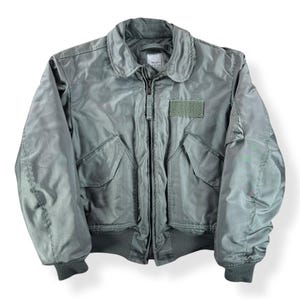 May include: A sage green bomber jacket with a zip-up front, two front pockets, and a patch on the chest. The jacket has a ribbed collar, cuffs, and hem. The jacket is made of a smooth material, possibly nylon.