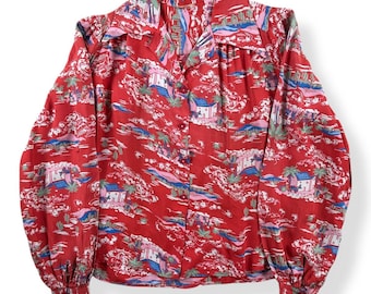 Pode incluir: Uma camisa vermelha com botões com uma estampa tropical com casas, palmeiras e nuvens. A camisa de manga comprida tem gola e punhos abotoados. O tecido tem um aspecto sedoso.
