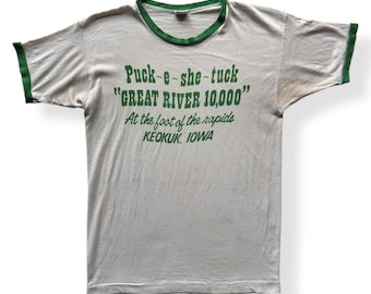 Könnte beinhalten: Ein weißes T-Shirt mit grünem Ringel. Das Shirt hat einen grünen Aufdruck mit der Aufschrift "Puck-e-she-tuck "GREAT RIVER 10,000" At the foot of the rapids KEOKUK, IOWA"