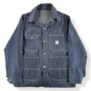 May include: A blue denim jacket with four front pockets, button closures, and white stitching. The jacket has a collar and a small label on one pocket. The sleeves are long, and the jacket appears to be a workwear style.