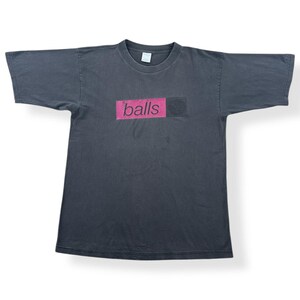 Pode incluir: T-shirt cinzento escuro de manga curta. A camisola apresenta a palavra "balls" num retângulo rosa, com um quadrado preto à direita. A camisola está estendida numa superfície branca.