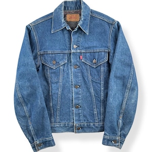May include: A classic blue denim jacket with a button-up front, two chest pockets, and a collar. The jacket features a red tag on the pocket and a brown label on the inside collar. The jacket is a medium wash denim.