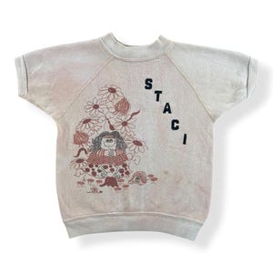 May include: A light beige short-sleeved sweatshirt featuring a graphic of a girl surrounded by flowers and mushrooms. The word "STACI" is printed vertically on the right side of the shirt in black letters. The sweatshirt has a crew neck and raglan sleeves.