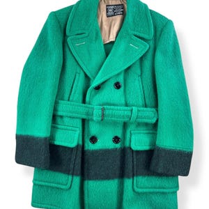 May include: A green wool coat with a double-breasted design, featuring black accents on the cuffs and a horizontal stripe across the midsection. The coat includes a matching belt and two front pockets. The coat has a label inside.