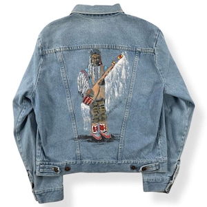 May include: Light blue denim jacket with a painted Native American figure on the back. The artwork features a detailed depiction of a person in traditional attire, holding a ceremonial object. The jacket has button closures and pockets.