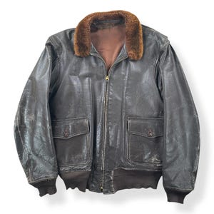 Peut inclure: Veste de bombardier en cuir marron avec un col en fourrure marron. La veste a deux poches avant, une fermeture éclair et des poignets et un ourlet côtelés. Le cuir montre des signes d'usure, lui donnant un aspect classique et vieilli.