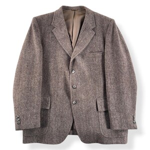 May include: A brown tweed blazer with a herringbone pattern. The jacket has a notched lapel, three buttons, and two front pockets. The sleeves have three buttons. The blazer is a classic style.