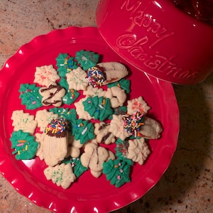May include: A red plate filled with assorted Christmas cookies, including tree-shaped and star-shaped cookies. Some cookies are decorated with green icing and sprinkles, while others are dipped in chocolate and topped with colorful sprinkles. A red bowl with "Merry Christmas" is in the background.