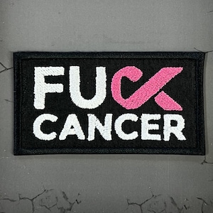 May include: Black rectangular patch with white embroidered text reading "FUCK CANCER". The pink cancer ribbon is incorporated into the word "FUCK". The patch is on a textured gray surface.