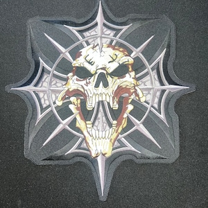 May include: A black patch featuring a detailed skull with an open mouth, set against a gray and black starburst design. The skull has a distressed, aged appearance with shades of brown and tan. The patch has a stitched border.