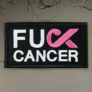May include: A rectangular black embroidered patch featuring the white text FUCK and the word CANCER in white below it. The letter C in the word FUCK is replaced by a pink awareness ribbon, with a black stitched border surrounding the entire design.