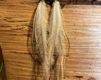 May include: A pair of long feather earrings. The earrings feature cream-coloured feathers with dark tips, suspended from gold-coloured metal hooks and decorative caps. The earrings are displayed against a textured, light brown wooden surface.