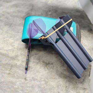 May include: A blue dartboard case with a rubber band holding a set of three darts. The darts have purple and blue fletching and a gold band around the barrel.