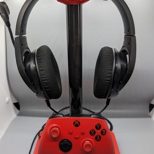 Peut inclure: Un support de casque de jeu rouge et noir avec une manette Xbox rouge sur la base. Le support a un poteau en métal noir et une base en plastique rouge. Le casque est noir avec des accents rouges. La manette a un logo Xbox noir et des boutons noirs.