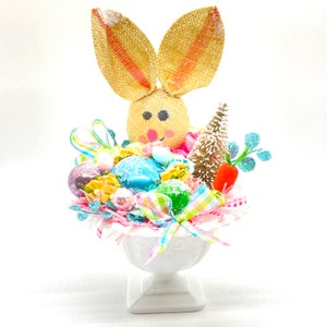 May include: A white ceramic pedestal vase filled with pastel Easter decorations, including a burlap bunny, pastel eggs, and a small pine tree.