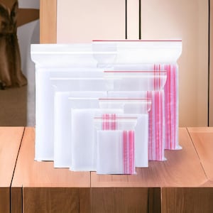 Pode incluir: Uma coleção de sacos de plástico transparentes com fecho zip em vários tamanhos. Os sacos têm uma faixa vermelha na parte superior e riscas rosa verticais. Apresentados numa superfície de madeira.