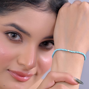 May include: A delicate bracelet featuring small, round, turquoise-colored beads strung together. A single silver-colored bead is interspersed within the turquoise beads. The bracelet is worn on a wrist, showcasing its simple elegance and vibrant color.