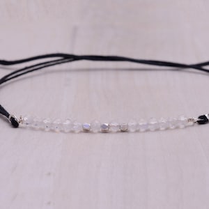 May include: A delicate bracelet with clear beads and silver accents strung on a thin, black cord. The cord is adjustable with sliding knots. The bracelet's design is minimalist and elegant, suitable for everyday wear. The beads are small and faceted.