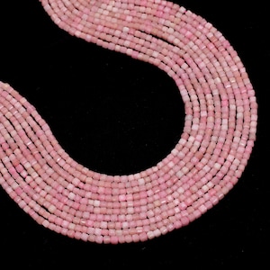 May include: Close-up of multiple strands of pink opal beads. The beads are small, square-shaped, and have a soft, pastel pink color. The strands are arranged in a flowing, curved pattern against a black background, showcasing the beads' texture and color.