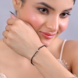 Op de afbeelding: Een delicate zwarte kralenarmband met een enkele witte kraal. De armband wordt om een pols gedragen en toont zijn eenvoudige elegantie. De kralen lijken klein en gelijkmatig verdeeld, waardoor een minimalistisch ontwerp ontstaat.