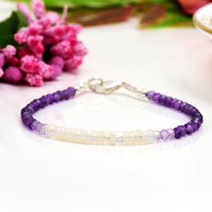 Könnte beinhalten: Ein zartes Armband aus Amethyst- und Opalperlen. Das Armband zeigt einen Farbverlauf von violetten Amethystperlen, die in der Mitte zu weißen Opalperlen übergehen. Das Armband wird mit einem silbernen Verschluss befestigt.