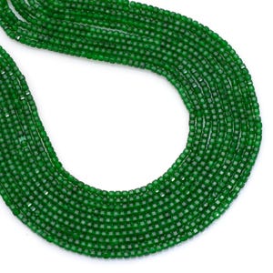 May include: Close-up of multiple strands of faceted, emerald green beads. The beads are small, square-cut, and strung together, creating a flowing, textured appearance. The vibrant green color is consistent throughout the strands, suggesting a gemstone or high-quality glass.