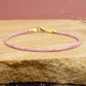 Peut inclure: Un bracelet délicat à perles roses avec un fermoir doré. Le bracelet est composé de petites perles de forme carrée. Le fond est une surface brun clair avec une toile de fond rose.