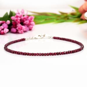 May include: A delicate bracelet made with small, square-shaped garnet beads in a deep red color. The bracelet is strung on a silver chain with a lobster clasp.