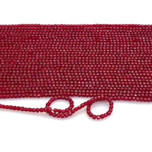 May include: A close-up view of a large collection of small, square-shaped, deep red beads. The beads are strung together, with a portion of the strand looped into a circle. The beads are evenly sized and have a faceted surface, creating a sparkling effect.