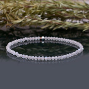 Puede incluir: Un delicado brazalete hecho con pequeñas cuentas de piedra lunar blanca iridiscente. El brazalete está enhebrado en una fina cadena plateada.