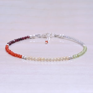 以下が含まれることがあります： 濃い赤からオレンジ、イエロー、そして明るいグリーンへとグラデーションになった宝石が並んだ、繊細なブレスレット。小さな丸いビーズで作られており、シルバーの留め具が付いています。