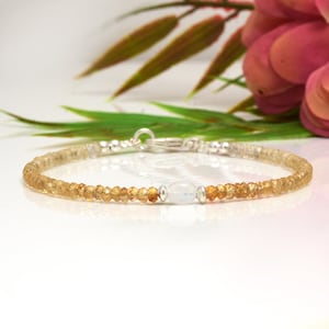 May include: A delicate bracelet made with tiny golden brown beads and a single white moonstone bead. The bracelet is secured with a silver clasp.