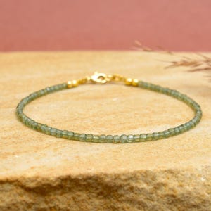 Puede incluir: Una delicada pulsera con pequeñas cuentas verdes claras ensartadas. La pulsera tiene un cierre y cuentas finales de color dorado. Las cuentas parecen ser de tamaño uniforme y la pulsera descansa sobre una superficie texturizada y bronceada.