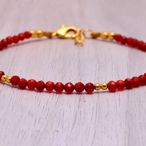 May include: A delicate bracelet featuring small, faceted red beads strung together. Gold-colored beads and a clasp accent the bracelet, creating a simple yet elegant design. The bracelet is approximately 7 inches in length.