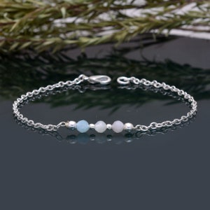 Könnte beinhalten: Ein zartes Armband aus silberner Kette mit drei pastellfarbenen Edelsteinperlen. Die Perlen sind hellblau, zartrosa und weiß.