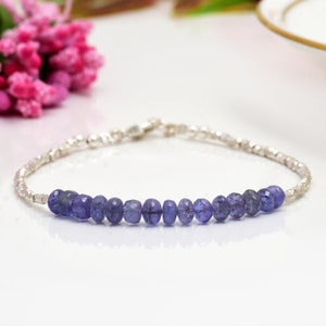 Könnte beinhalten: Ein silbernes Armband mit einer Reihe kleiner, runder, lilafarbener Edelsteine. Das Armband liegt auf einer weißen Oberfläche.