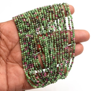 Peut inclure: Gros plan de plusieurs brins de petites perles à facettes vertes, roses et noires. Les perles sont enfilées sur un fil rouge. Les perles sont tenues dans une main sur fond blanc. Idéal pour la création de bijoux.