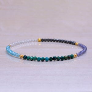 May include: A multi-colored beaded bracelet with a gradient of light blue, green, gray, and purple beads. The bracelet is made with small, round beads and has gold accents.