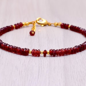 Puede incluir: Una pulsera de piedras preciosas rojas con detalles dorados. La pulsera presenta pequeñas cuentas rojas facetadas ensartadas, intercaladas con cuentas doradas y un cierre dorado. La pulsera se muestra sobre una superficie clara.