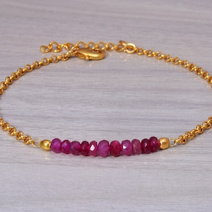 May include: A gold-colored chain bracelet featuring a row of faceted, deep red ruby beads. The bracelet has a delicate, textured chain and a simple clasp. The gemstones are the focal point, set against a neutral background.