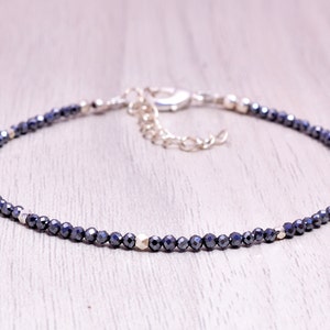 Könnte beinhalten: Ein zartes Armband mit kleinen, facettierten, dunkelblauen Perlen. Das Armband hat silberfarbene Akzente und einen Verschluss. Die verstellbare Kette ermöglicht eine bequeme Passform. Dieses Schmuckstück ist für den täglichen Gebrauch geeignet.